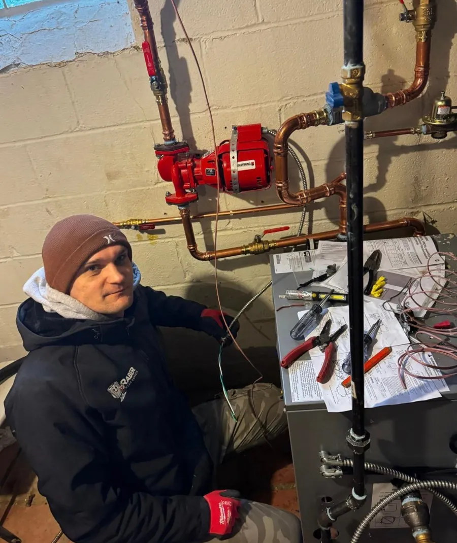 Raber technician in a branded jacket wiring the control circuit on a boiler beside a red circulator pump and copper piping